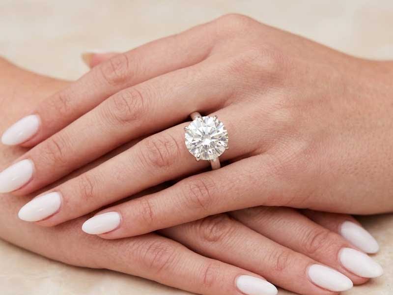 A hand with manicured nails wears a large, round diamond solitaire ring on the ring finger.