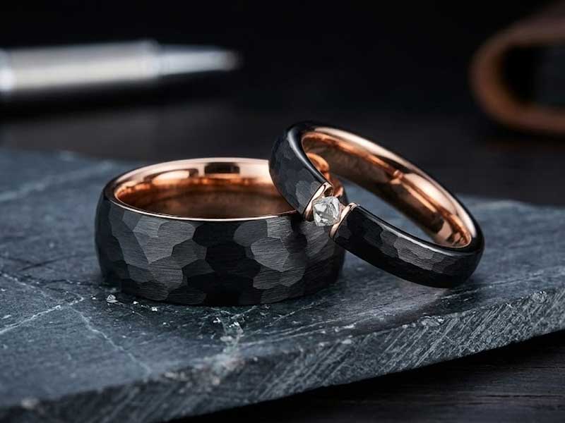 Two black and rose gold rings rest on a dark textured stone surface; one ring has a diamond solitaire setting.