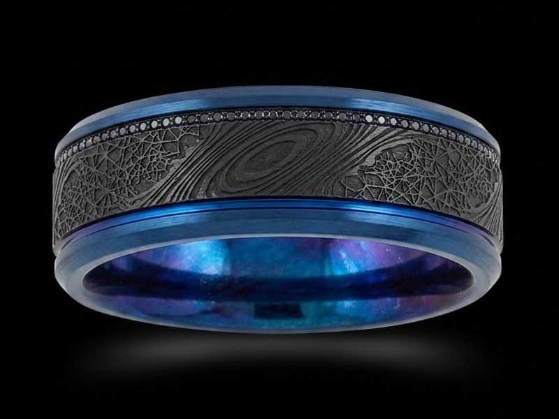 A black and blue ring with a textured outer band featuring an engraved planetary design and small black stones along the edge, set against a black background.