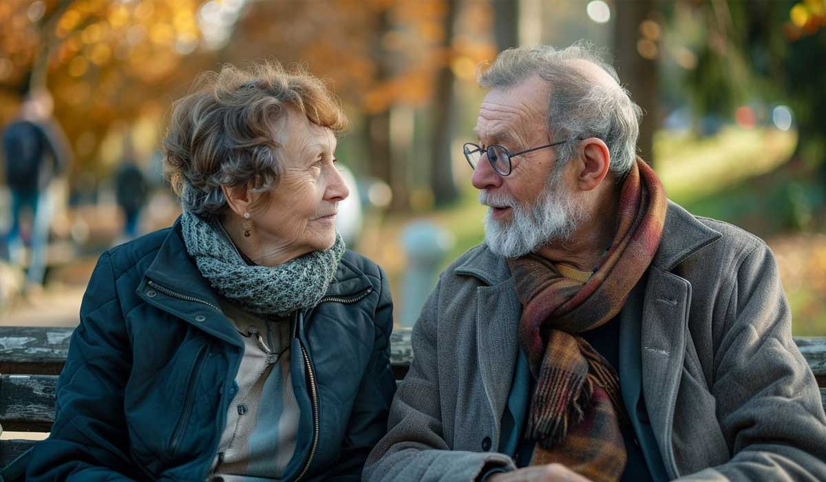 An elderly man and woman sit on a bench outdoors in autumn, facing each other and talking. Both are wearing jackets and scarves, with trees and people blurred in the background.