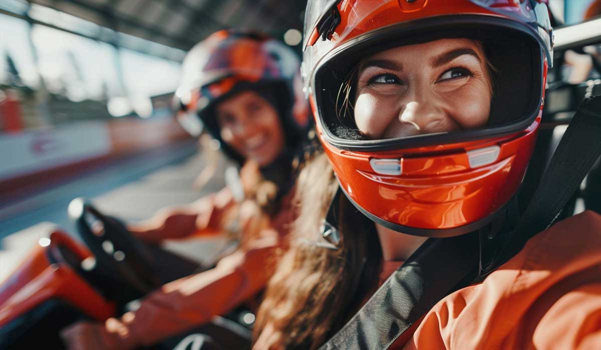 Two women wearing helmets and racing suits sit in go-karts on an indoor track, smiling and looking ahead.