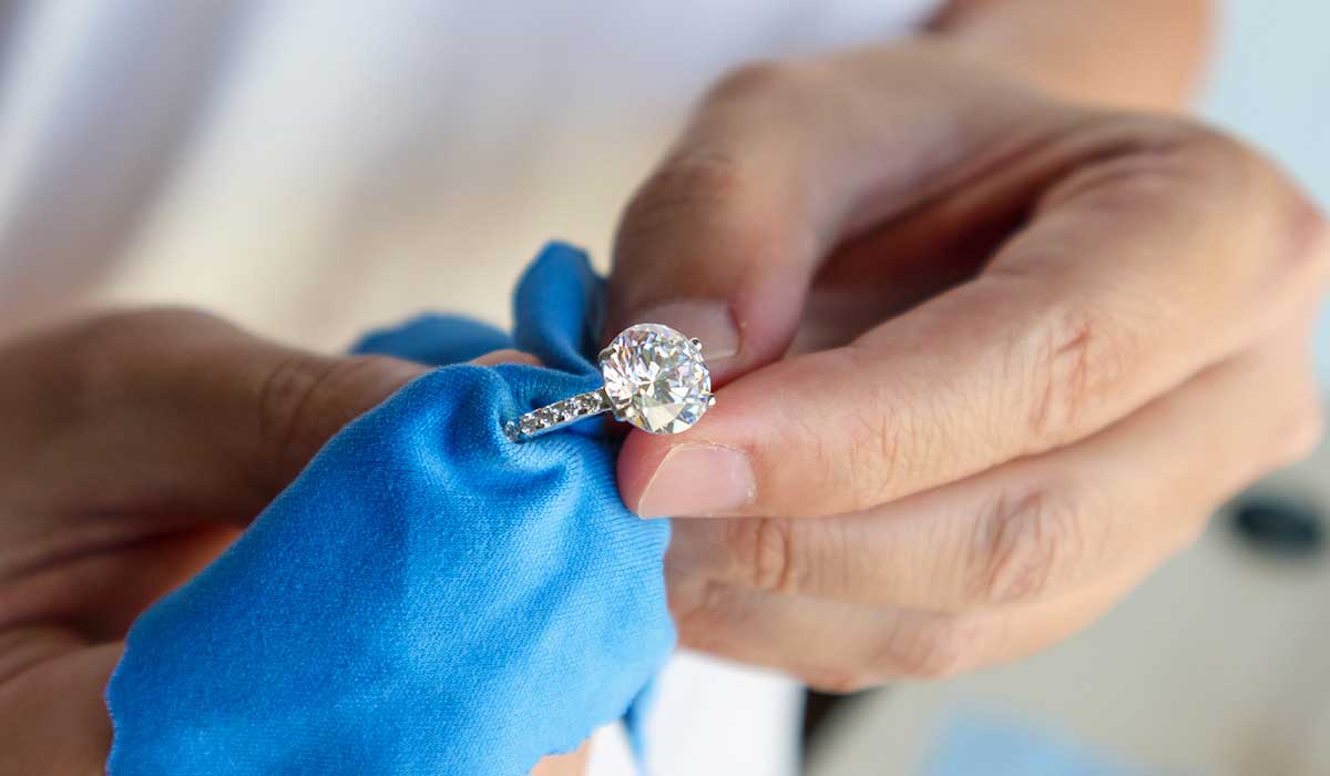 A person polishes a diamond ring with a blue cloth, focusing on the large central gemstone and the band.