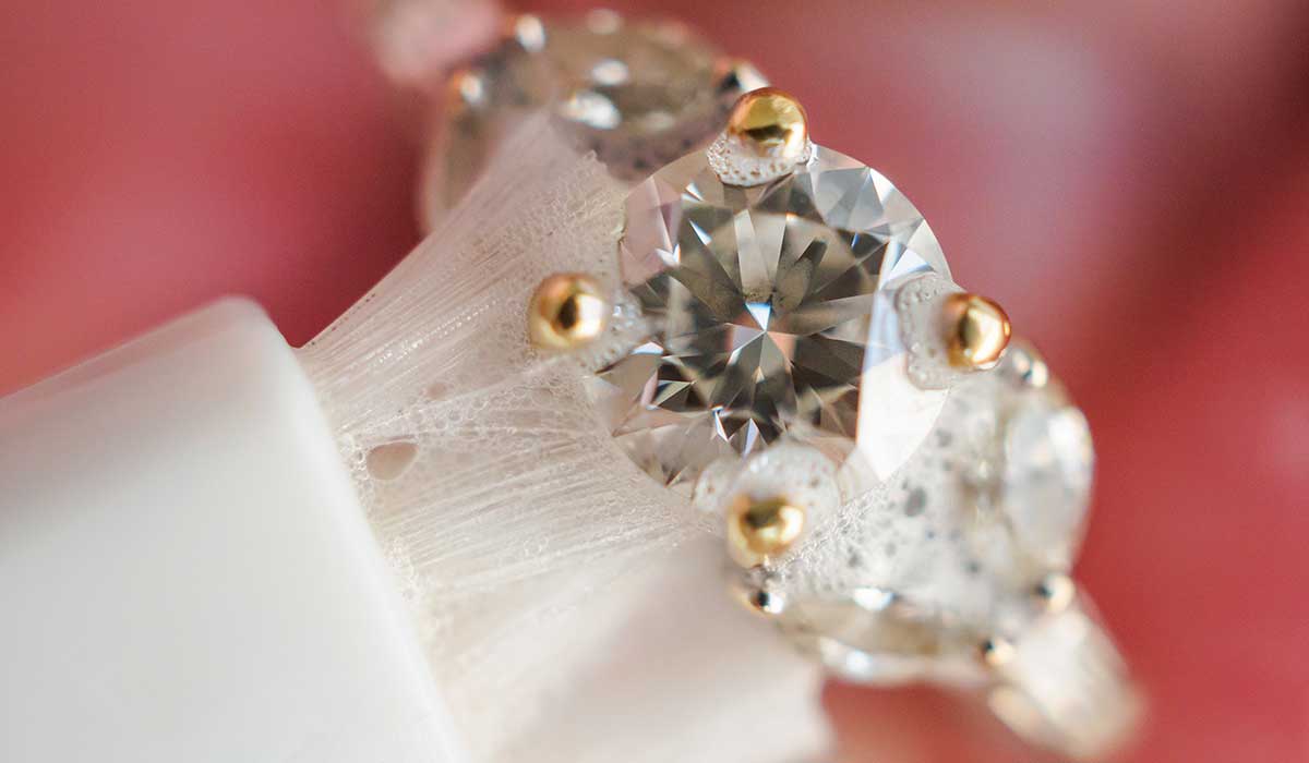 Close-up of a diamond ring with a large central diamond set in gold prongs, surrounded by smaller diamonds on a blurred pink background.