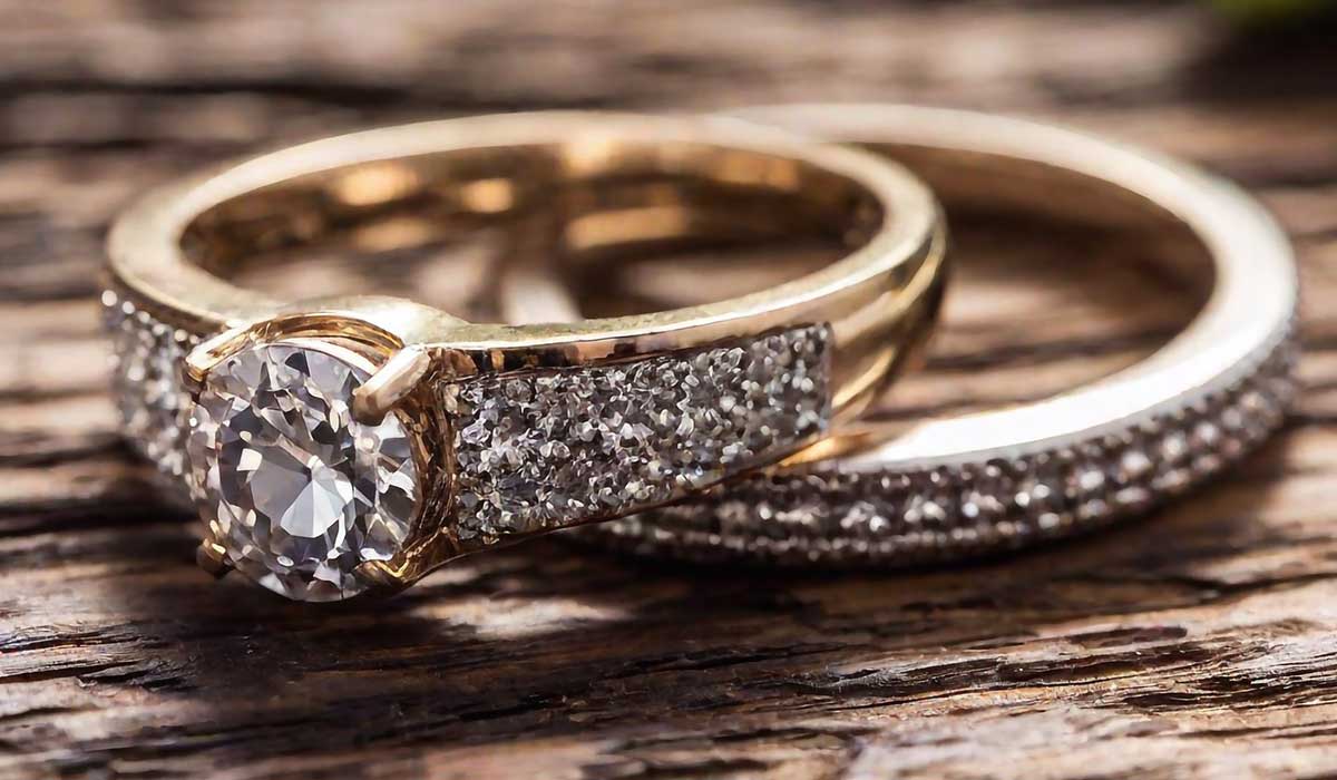 Two gold rings rest on a wooden surface; one features a large round diamond in a prong setting with smaller stones, and the other is a matching diamond-studded band.