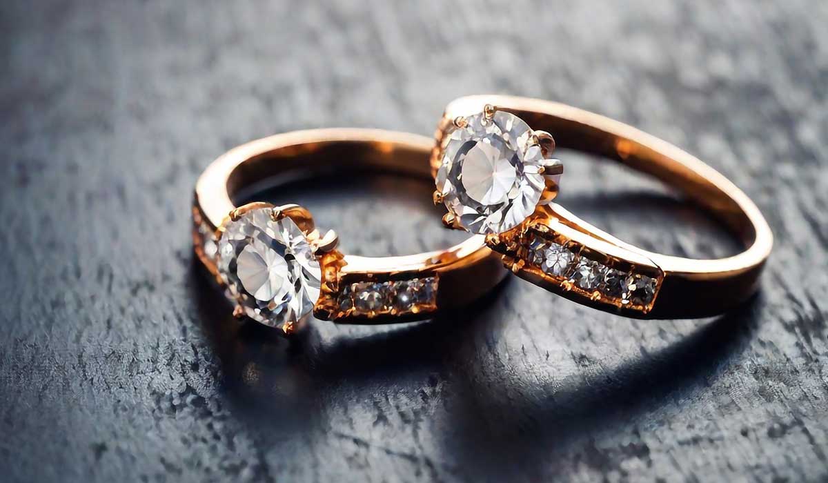Two gold rings with round diamonds and small stones on their bands are placed on a dark, textured surface.