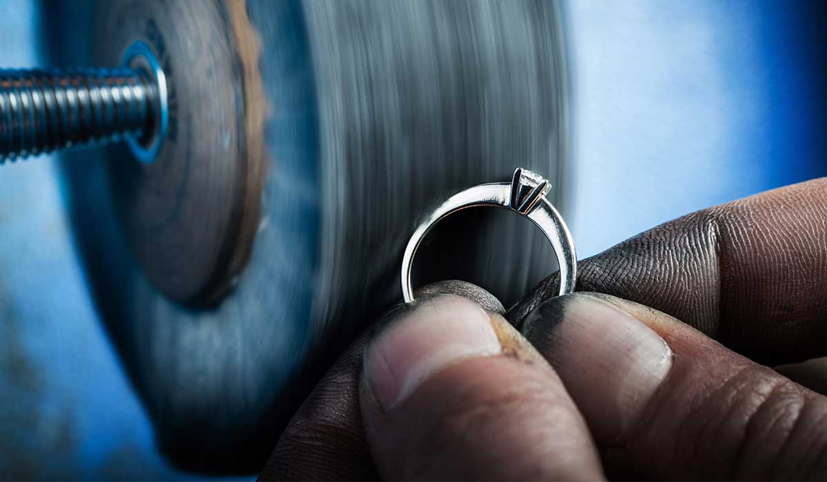 A person holds a diamond ring close to a spinning polishing machine, refining the ring’s surface.