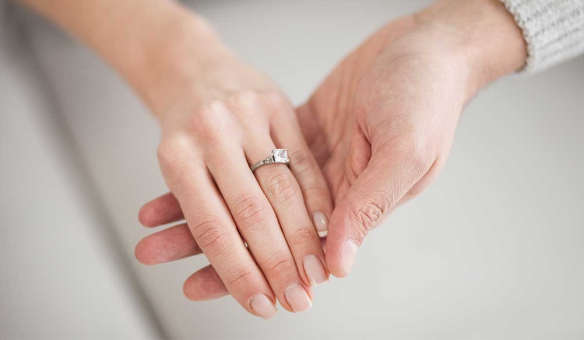 A close-up of one hand wearing a diamond ring being gently held by another hand against a light background.