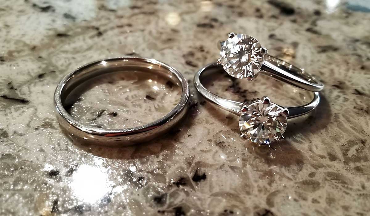 A plain metal wedding band next to two diamond solitaire engagement rings resting on a speckled granite surface.