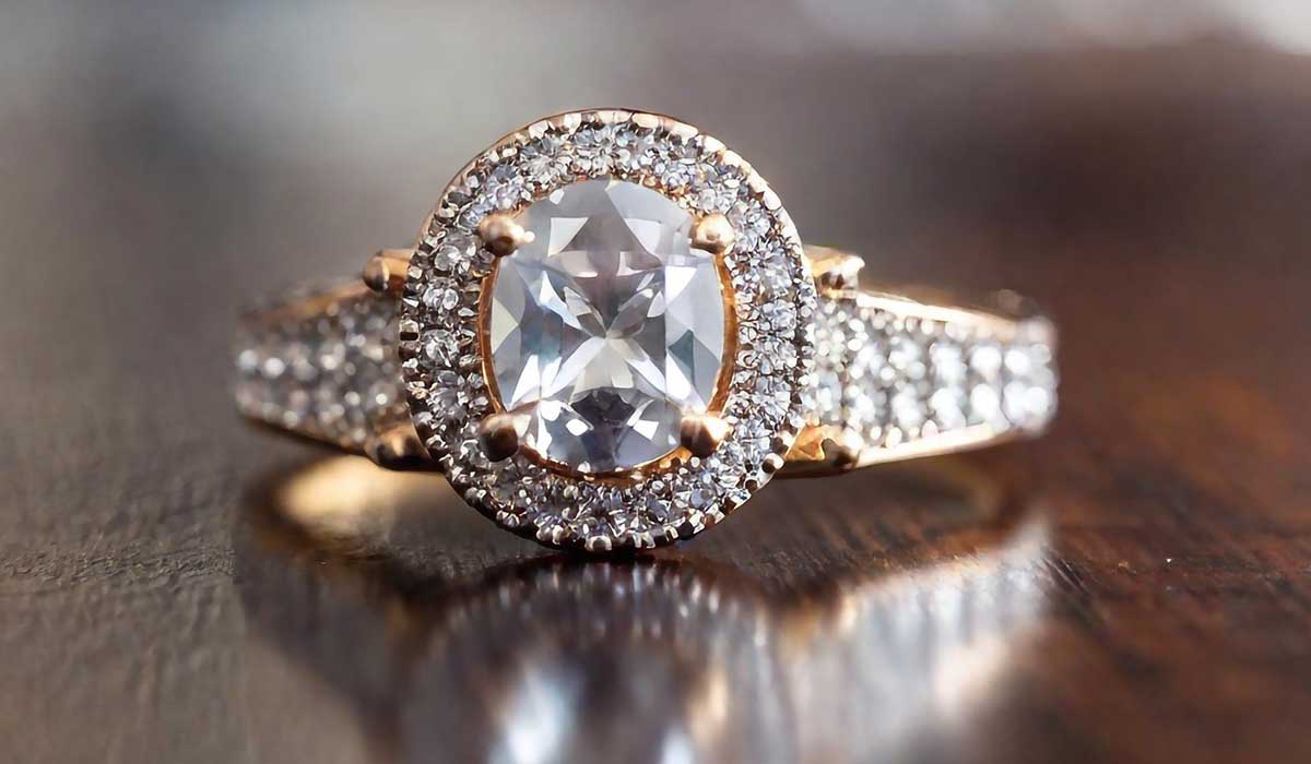 A gold ring with a large cushion-cut center diamond, surrounded by smaller diamonds, is displayed on a wooden surface.