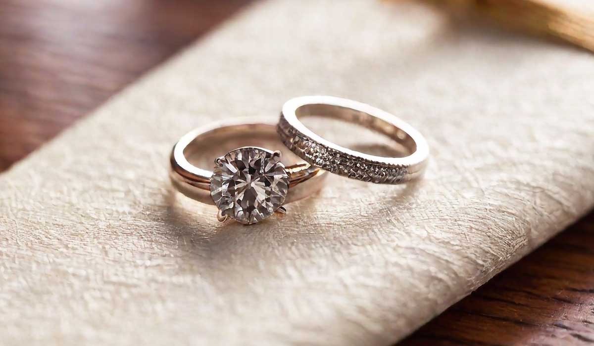Two gold rings on a textured surface; one is a solitaire diamond engagement ring, the other is a wedding band with small embedded diamonds.
