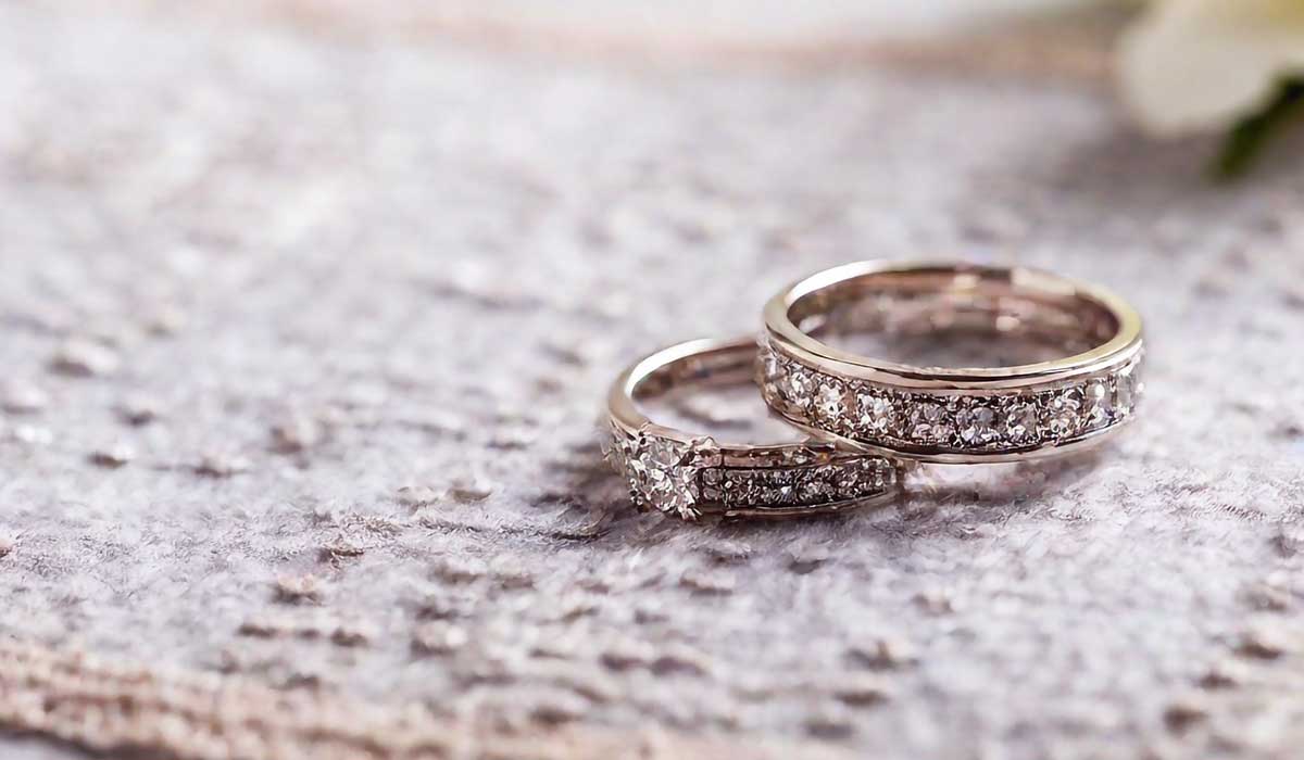 Two diamond-studded gold rings rest on a textured, light-colored fabric surface, with a blurred white flower visible in the background.