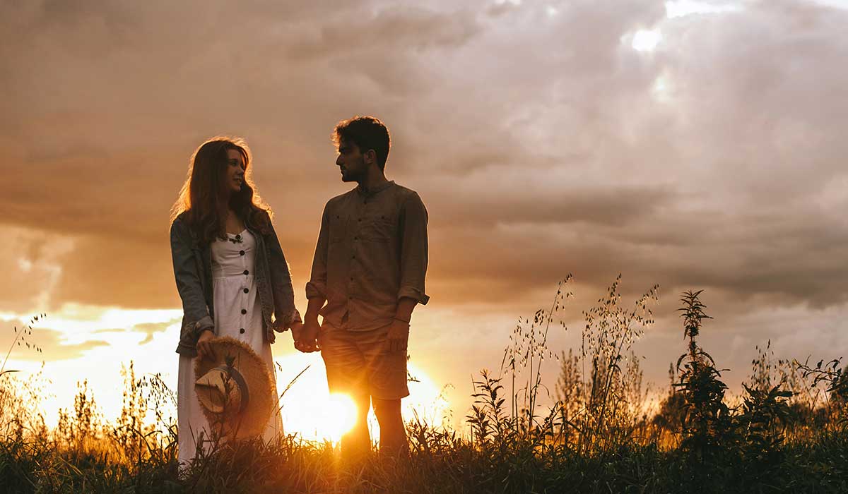 A woman and man stand in tall grass at sunset, holding hands and looking at each other under a cloudy sky.
