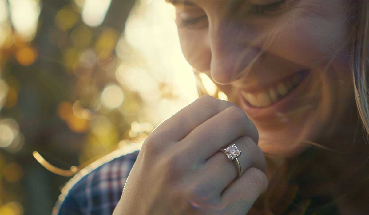 A close-up of a smiling woman outdoors holding her hand to her face, showing an engagement ring on her finger in soft sunlight.