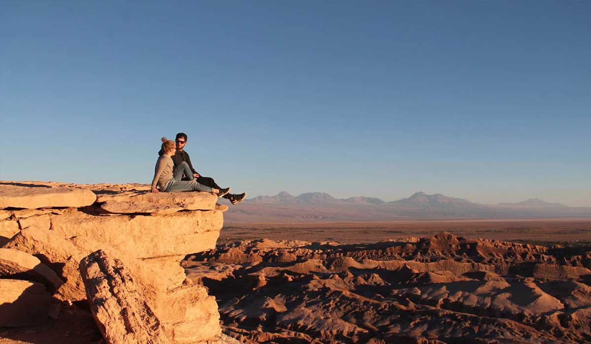 Two people sit on the edge of a rocky cliff overlooking a vast desert landscape under a clear blue sky.