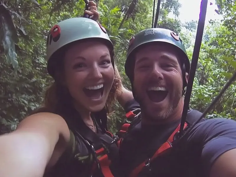 Two people wearing helmets and harnesses are smiling widely while ziplining in a forested area.