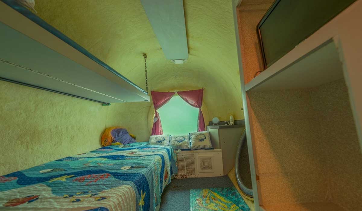 Small room with yellow curved walls, a single bed with a colorful quilt, shelves, a window with red curtains, and a TV on a shelf.