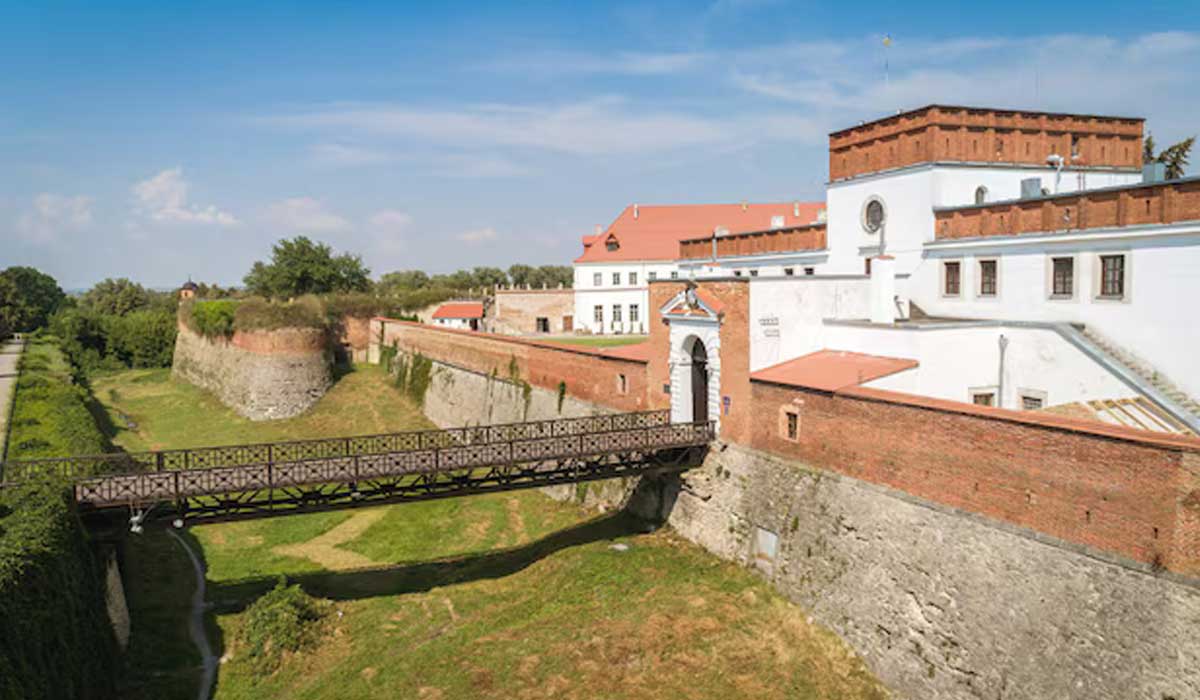 A historic fortress with white walls, a red-roofed tower, and a wooden bridge crossing a grassy moat on a sunny day.