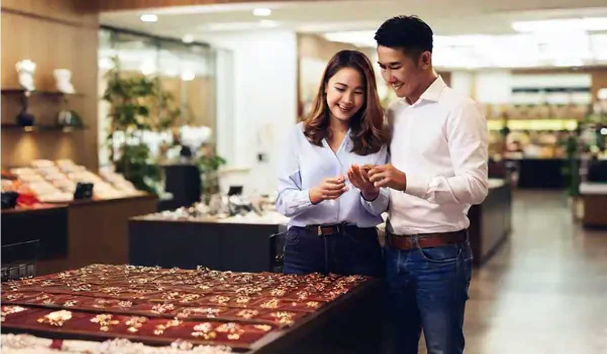 A man and woman stand together in a jewelry store, looking at rings displayed on a counter.