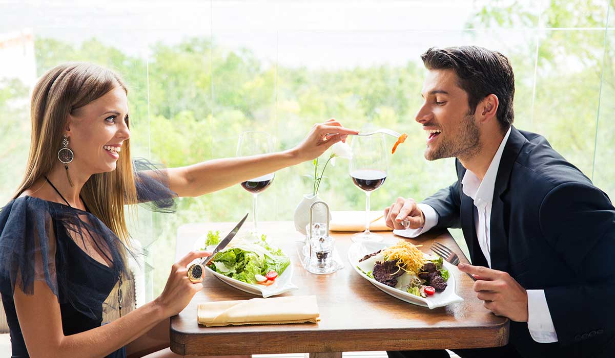 A woman and a man sit at a restaurant table eating pasta and salad, with wine glasses in front of them. The woman is feeding the man with a fork.