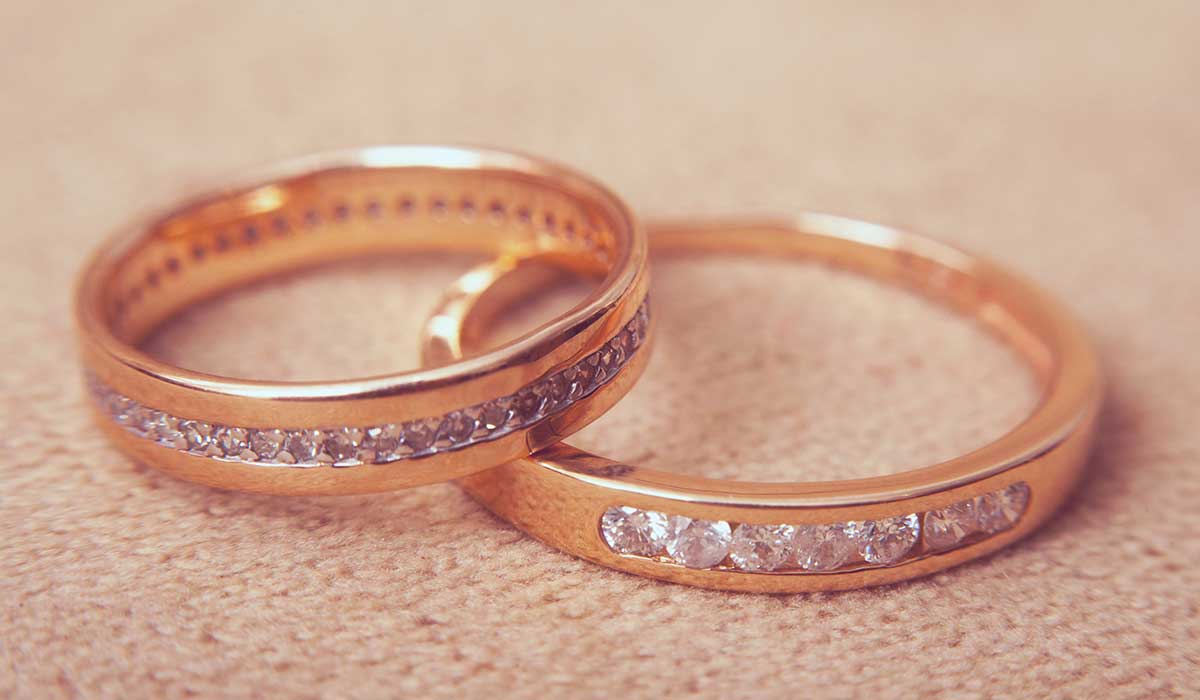Two gold rings with small diamonds set in a row, lying on a light brown textured surface.