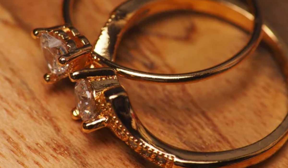 Two gold rings with clear gemstones rest on a wooden surface, with one ring featuring a single stone and the other a detailed band.