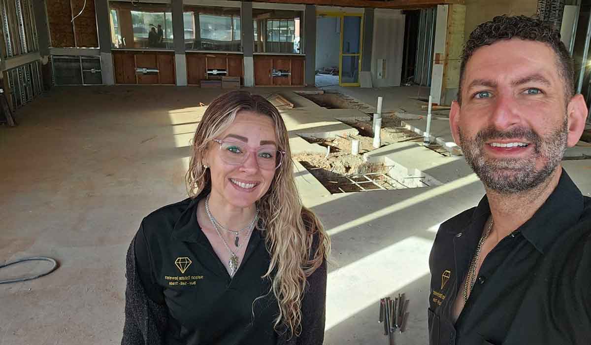 Two people in black shirts stand inside a large, unfinished room with exposed flooring, piping, and construction materials.