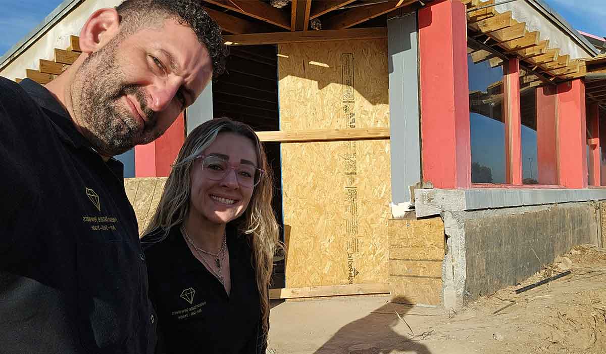 Two people stand in front of a partially constructed building with plywood covering the entrance and exposed framing visible.