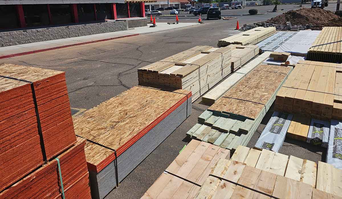 Stacks of lumber and plywood are arranged on the ground in a parking lot near a building, with orange cones and dirt piles in the background.