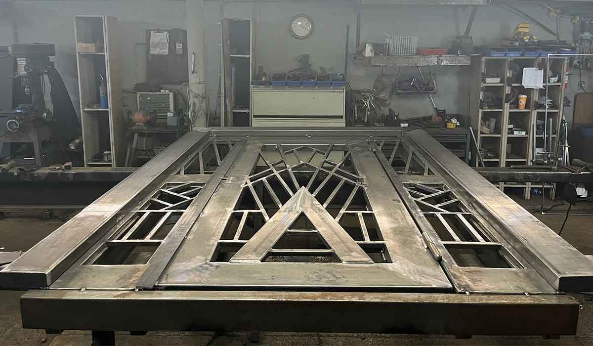 Large geometric metal gate in a workshop, surrounded by industrial tools, shelves, and equipment.