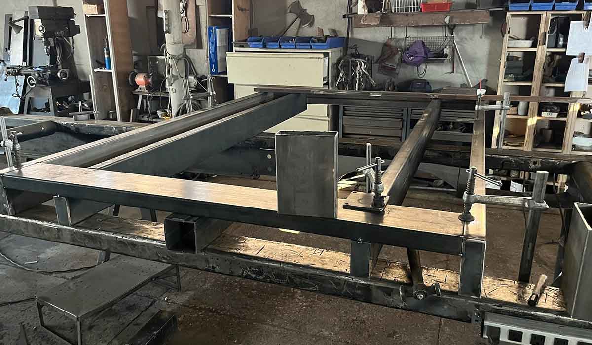 Metal frame under construction with rectangular beams and clamps on a workbench in an industrial workshop. Tools, shelves, and equipment are visible in the background.