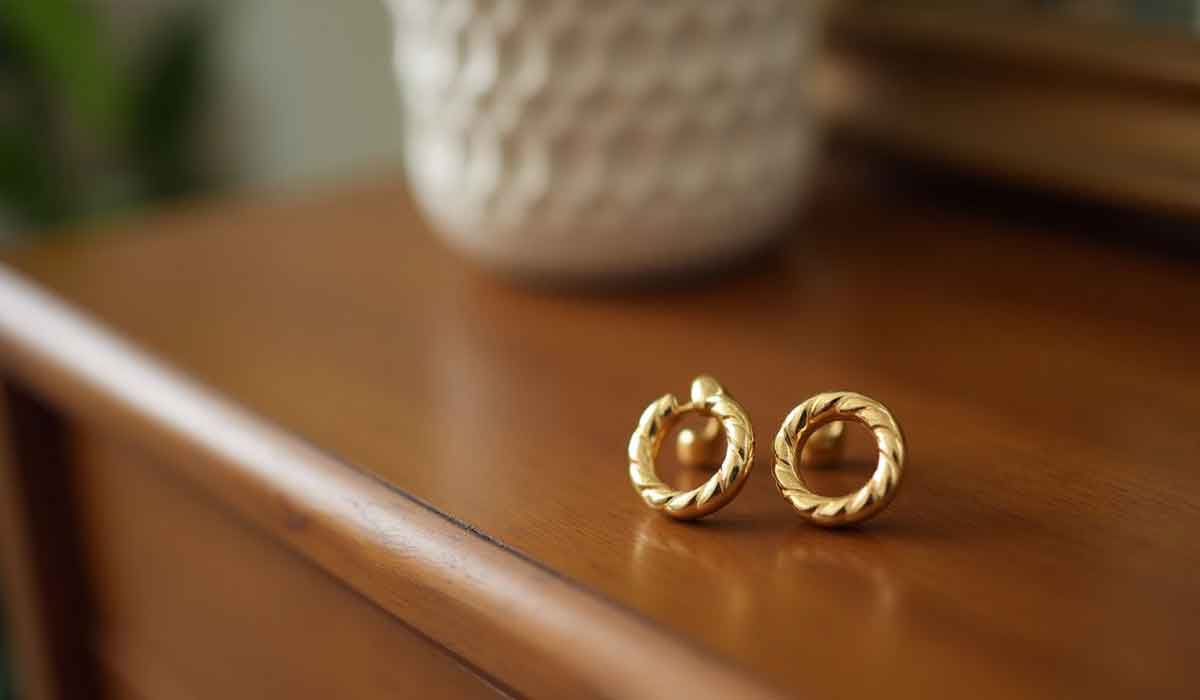 A pair of gold, twisted hoop earrings rests on a polished wooden surface, with a textured white vase blurred in the background.
