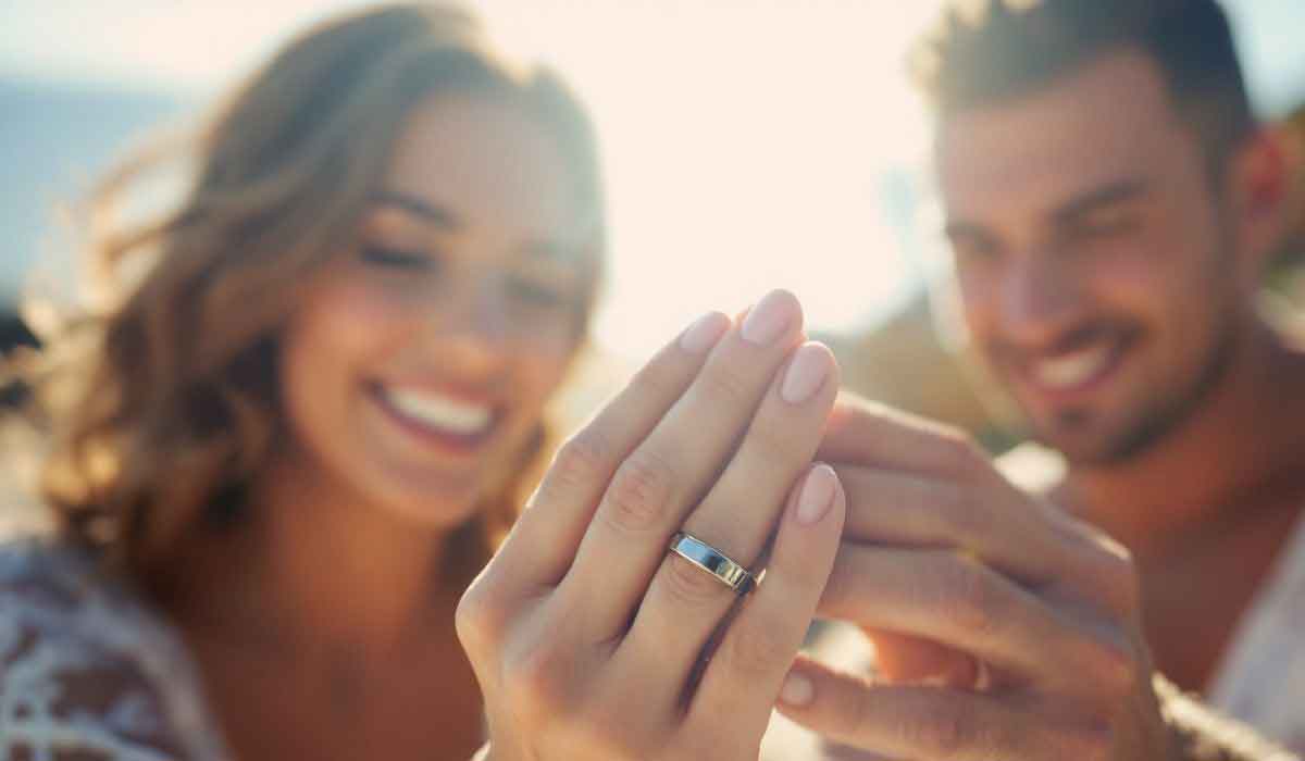 A woman shows an engagement ring on her hand while smiling next to a man outdoors in bright sunlight.
