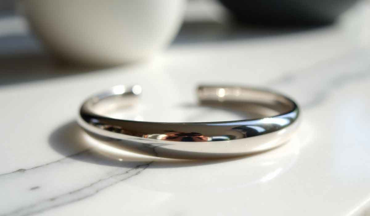 A silver cuff bracelet rests on a white marble surface with natural light reflecting off its polished finish.