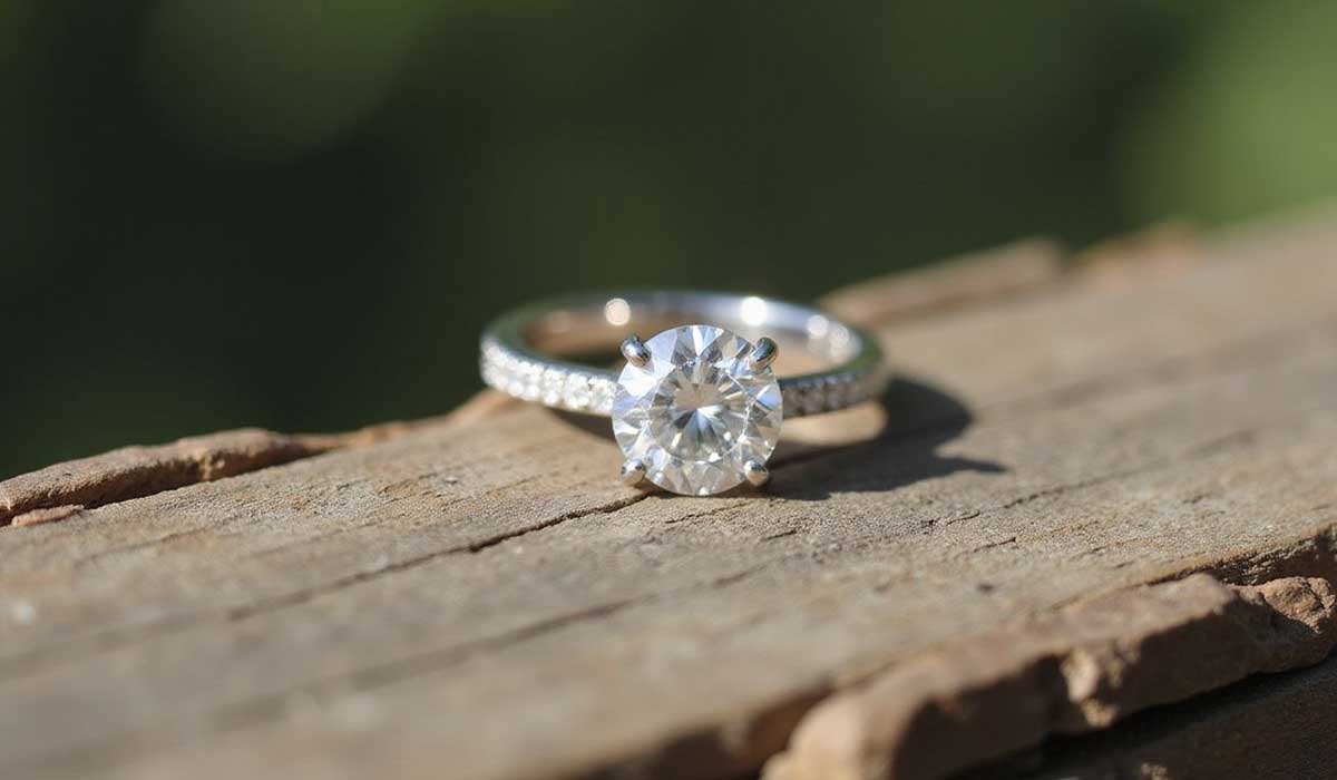 A diamond engagement ring with a round center stone and a band featuring small diamonds, resting on a weathered wooden surface.
