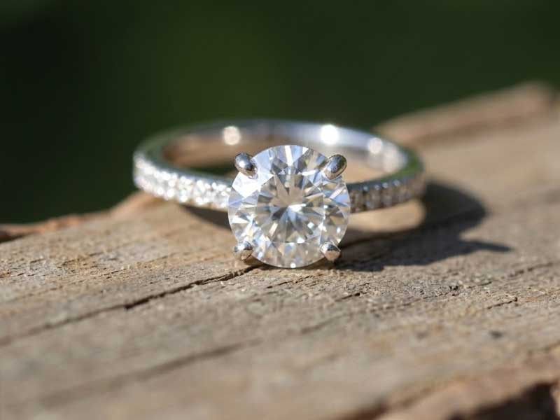 A silver ring with a large round diamond and smaller diamonds on the band sits on a weathered wooden surface.