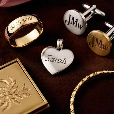 A gold ring engraved with "06.15.2019," a silver heart pendant engraved with "Sarah," two monogrammed cufflinks, a gold bracelet, and a gold engraved case on a brown surface.