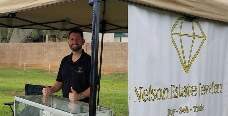 A man stands behind a glass display case at an outdoor booth labeled "Nelson Estate Jewelry" with a diamond logo, giving two thumbs up.