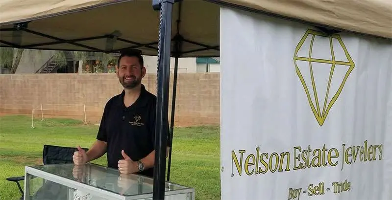 A man stands behind a glass display case at an outdoor booth labeled "Nelson Estate Jewelry" with a diamond logo, giving two thumbs up.