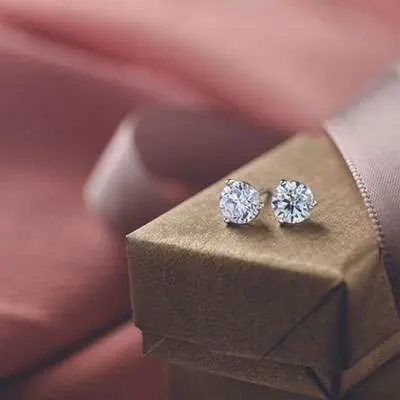 A pair of diamond stud earrings rests on a small, wrapped gift box with a pink ribbon in the background.