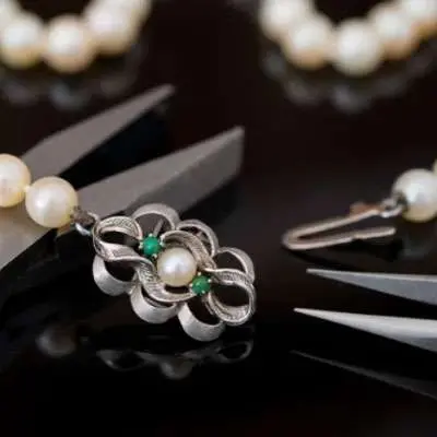 Close-up of jewelry making tools working on a silver and pearl pendant with green bead accents, surrounded by pearl necklaces on a dark surface.
