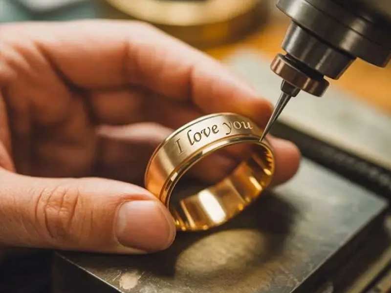 A close-up of a hand holding a gold ring while a machine engraves the words “I love you” on its surface.