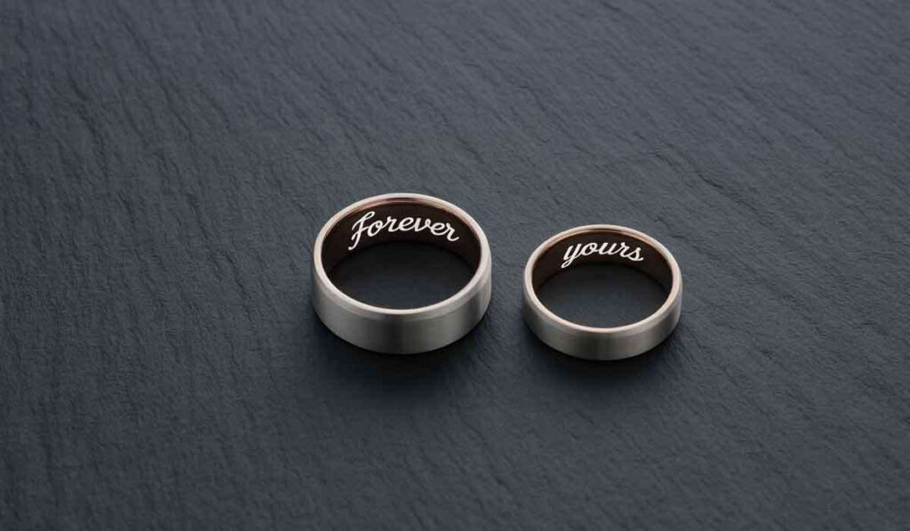 Two silver rings placed on a dark surface; one ring is engraved with "forever" and the other with "yours" on their inner bands.