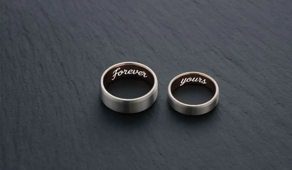 Two silver rings placed on a dark surface; one ring is engraved with "forever" and the other with "yours" on their inner bands.