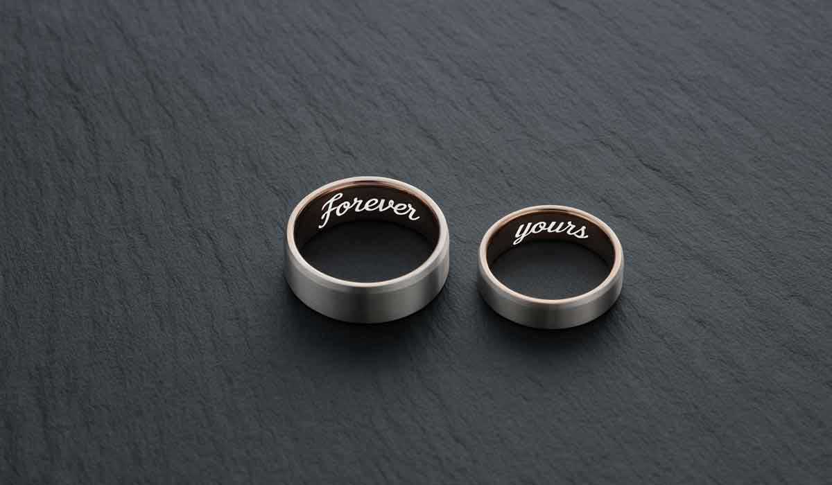 Two silver rings on a dark surface; one ring is engraved with “Forever” and the other with “yours” on the inside.