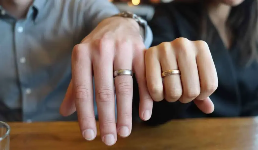 Two people sit side by side at a table, each displaying a hand with a matching ring on their ring finger.