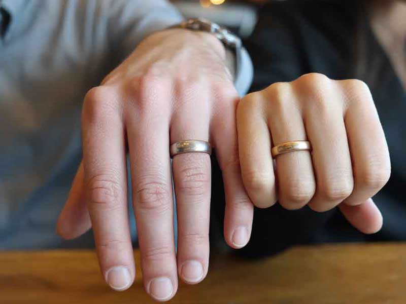 Two people sit side by side showing their left hands with wedding rings on their ring fingers, resting on a wooden surface.
