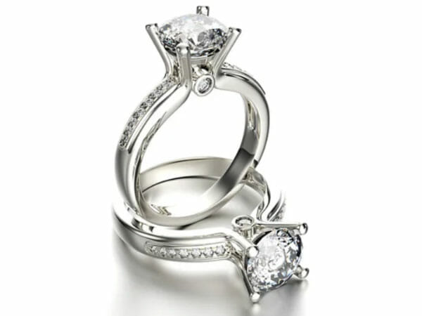 Two silver engagement rings with round-cut diamonds set in raised prongs, each band featuring smaller embedded diamonds, displayed on a white background.