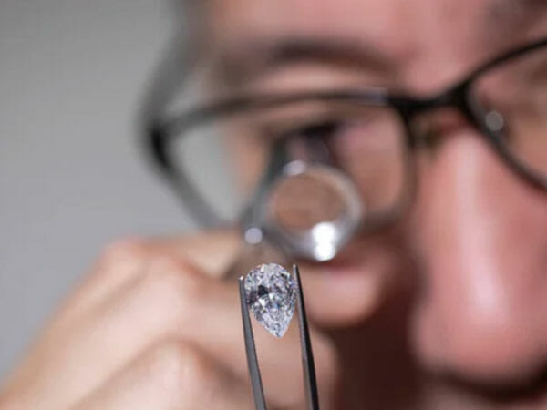 A person examines a pear-shaped diamond with tweezers and a jeweler’s loupe.