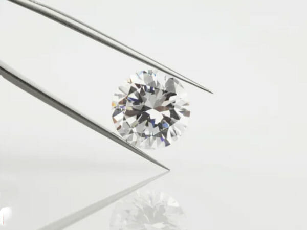 A pair of tweezers holds a round, faceted diamond against a plain white background, with the diamond’s reflection visible below.