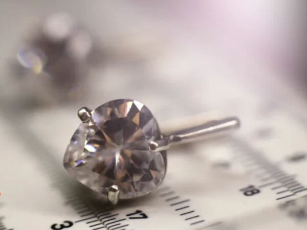 A heart-shaped diamond held by metal prongs rests on a ruler, with blurred background elements and soft lighting.